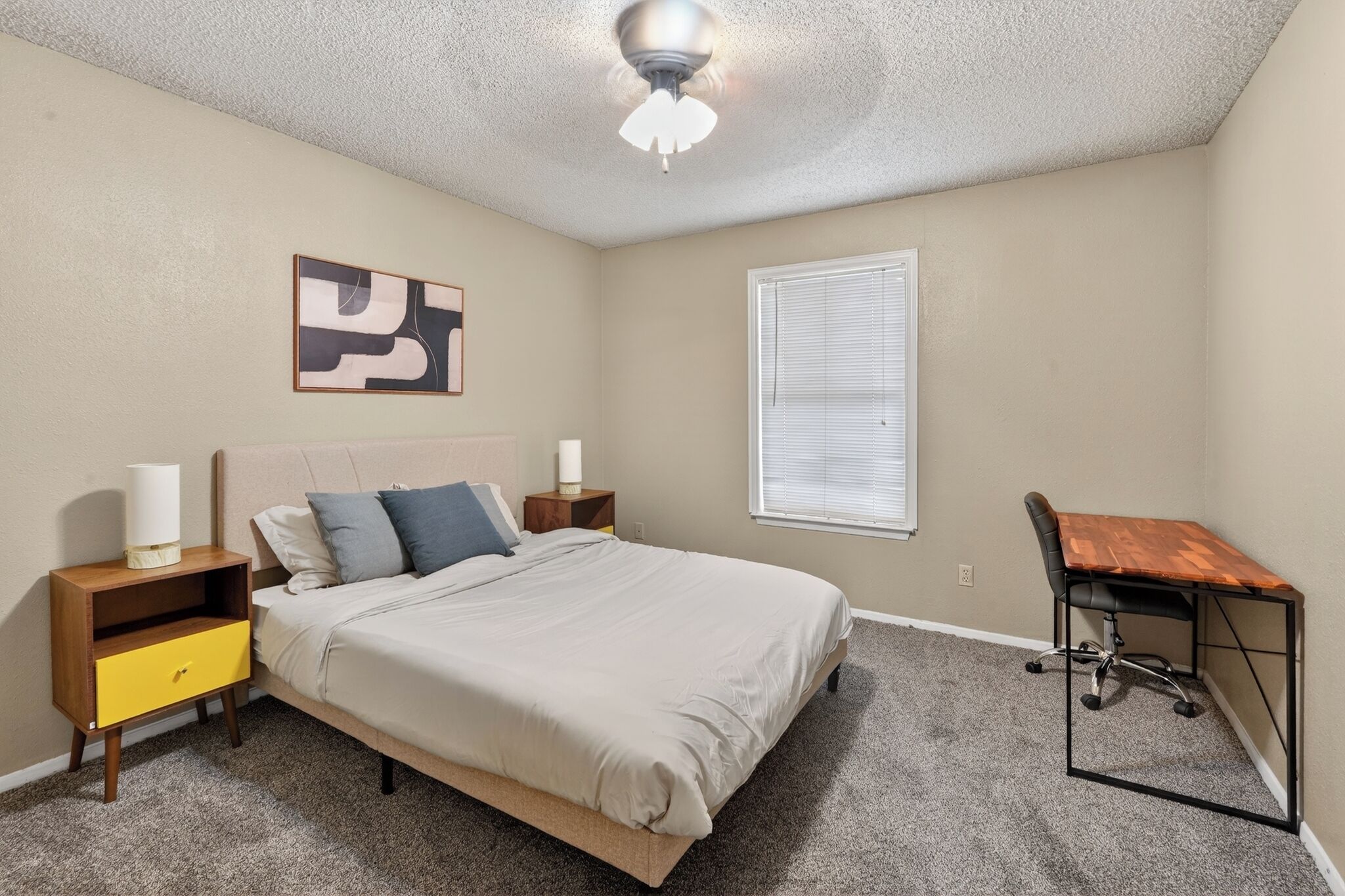 A bedroom with a bed, bedside tables, a desk, a ceiling fan, and a window.