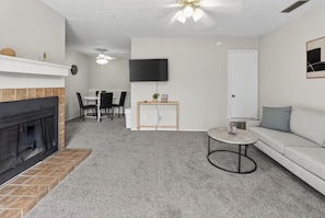 A living room with a fireplace, a television mounted on the wall, a sofa, a small round table, and a dining area with chairs. - Updated 2BR with great natural light (Lubbock)