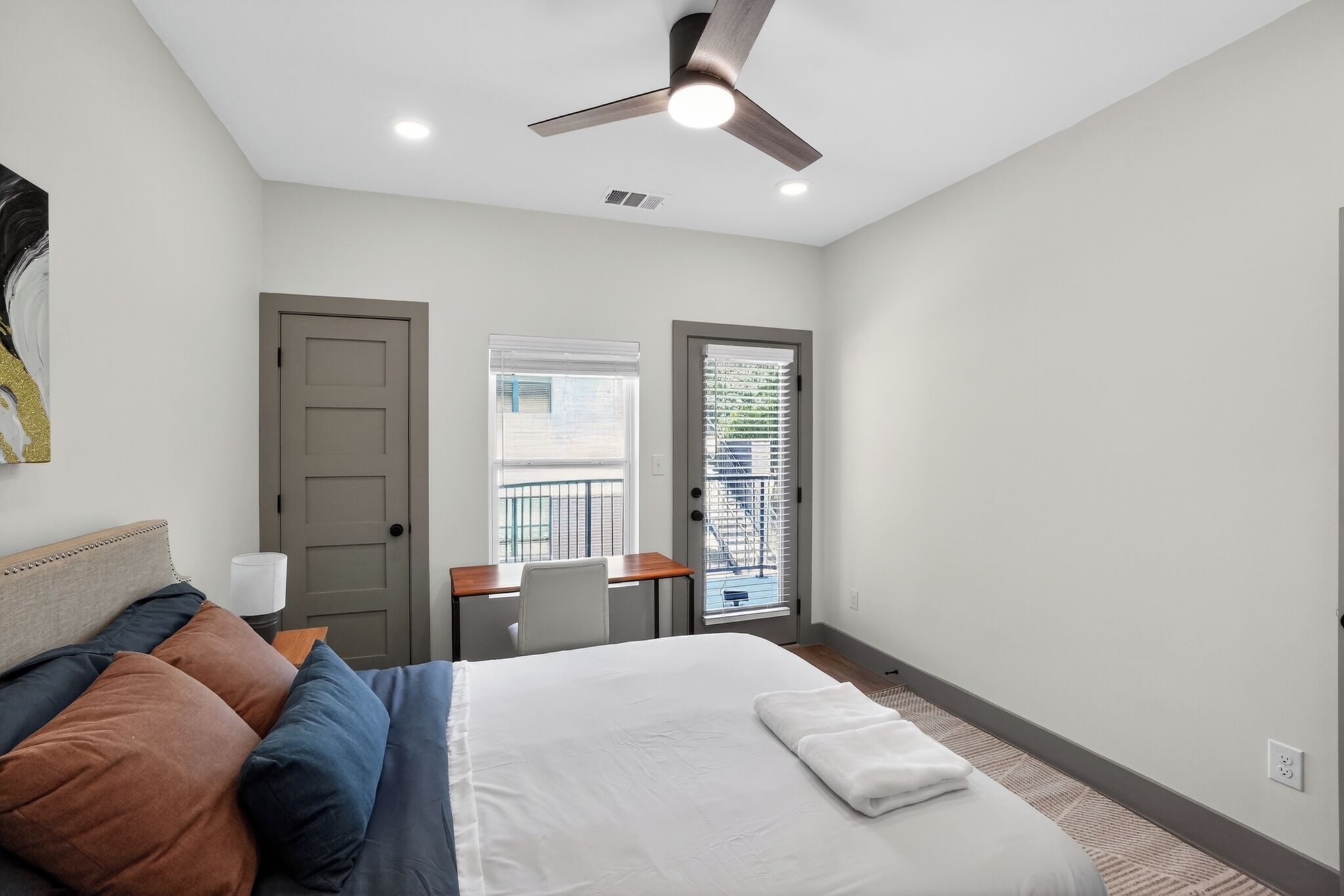 A bedroom with a bed, a desk, a chair, a ceiling fan, and a window with blinds.
