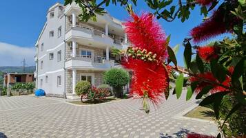 A multi-story residential building with a paved driveway, surrounded by greenery and red flowering plants.