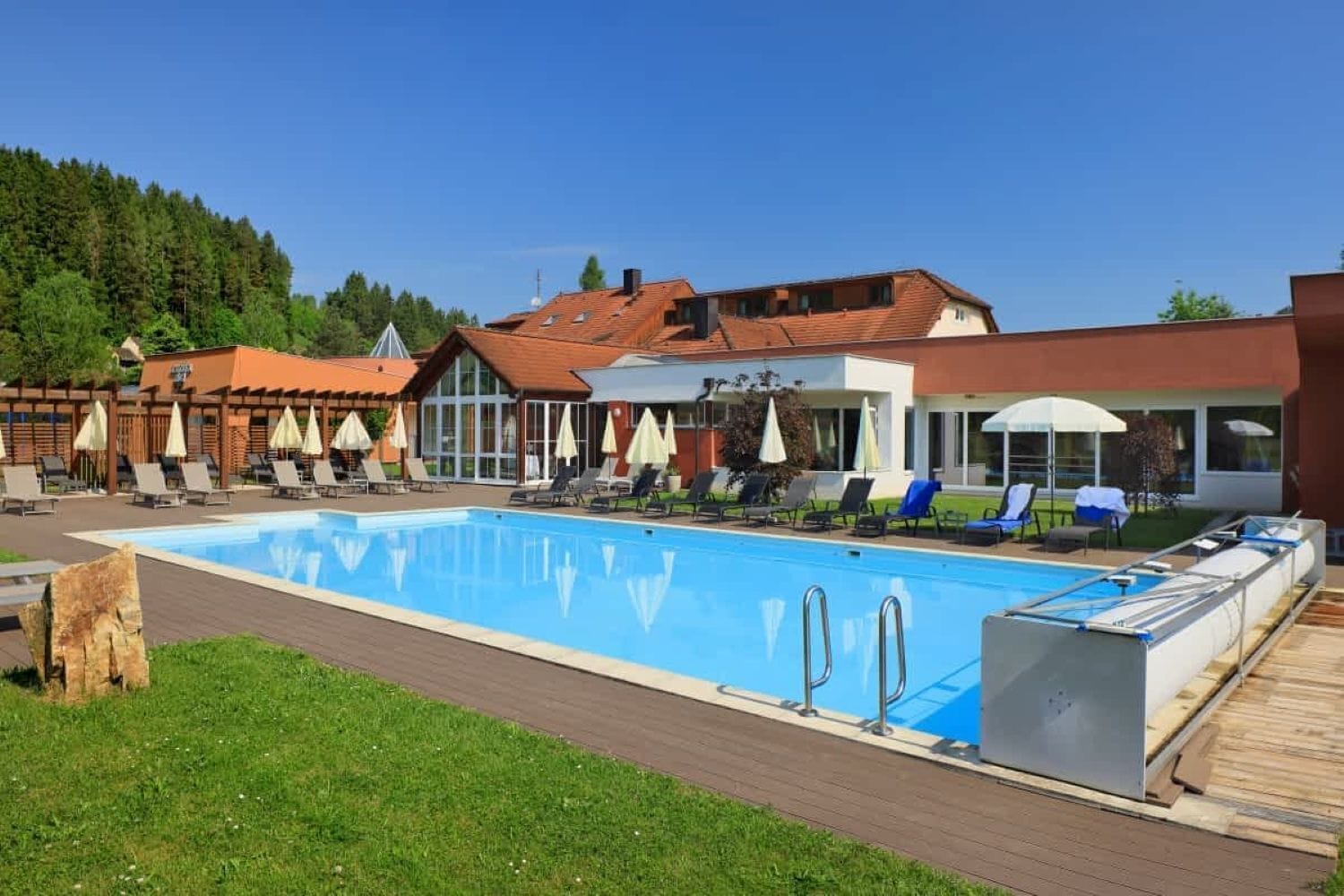 A swimming pool with lounge chairs and umbrellas, surrounded by buildings with red roofs and a forested hill in the background.