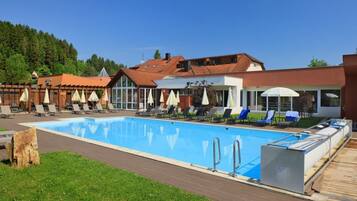 A swimming pool with lounge chairs and umbrellas, surrounded by buildings with red roofs and a forested hill in the background.