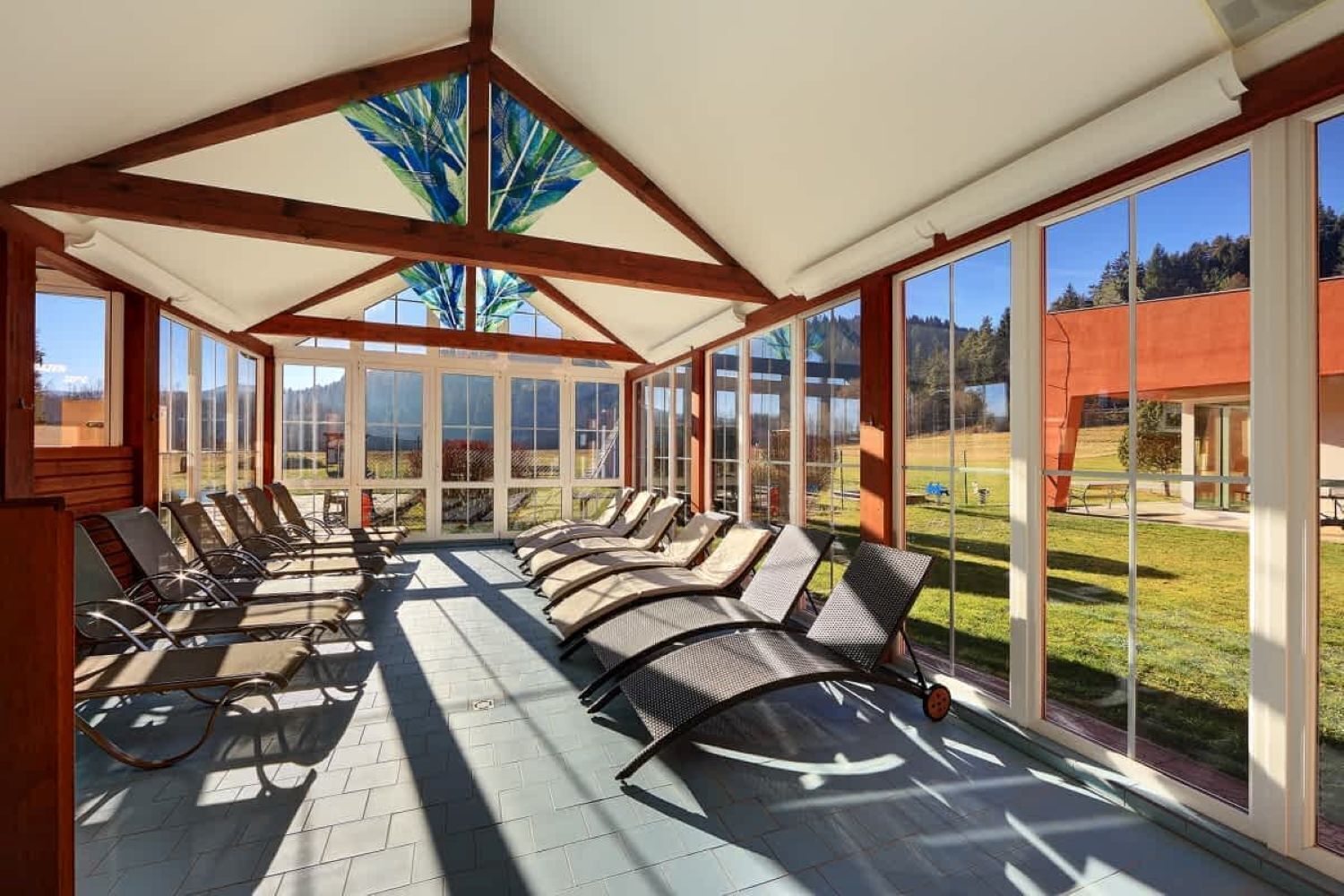 A sunroom with large windows, a ceiling with wooden beams, and lounge chairs arranged for sunbathing.