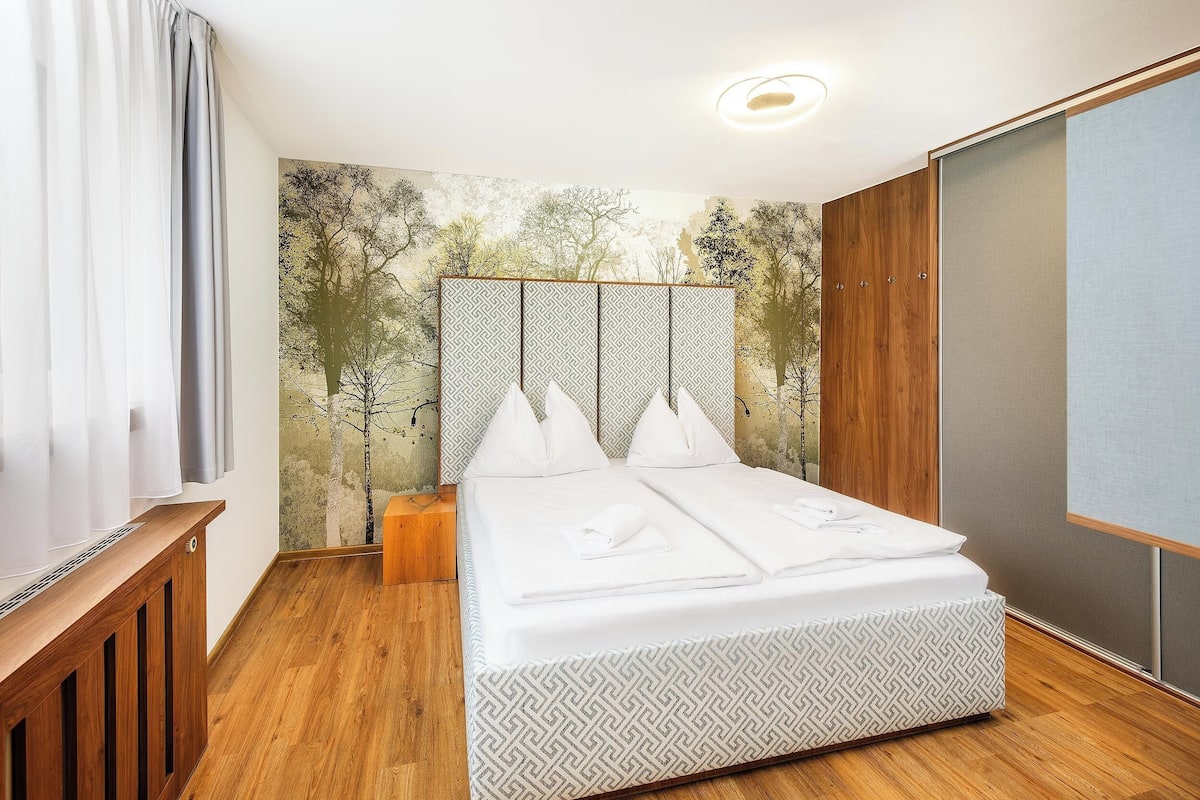 A neatly made bed with a patterned bed skirt, a headboard with a geometric design, and a wall with a nature-themed mural.