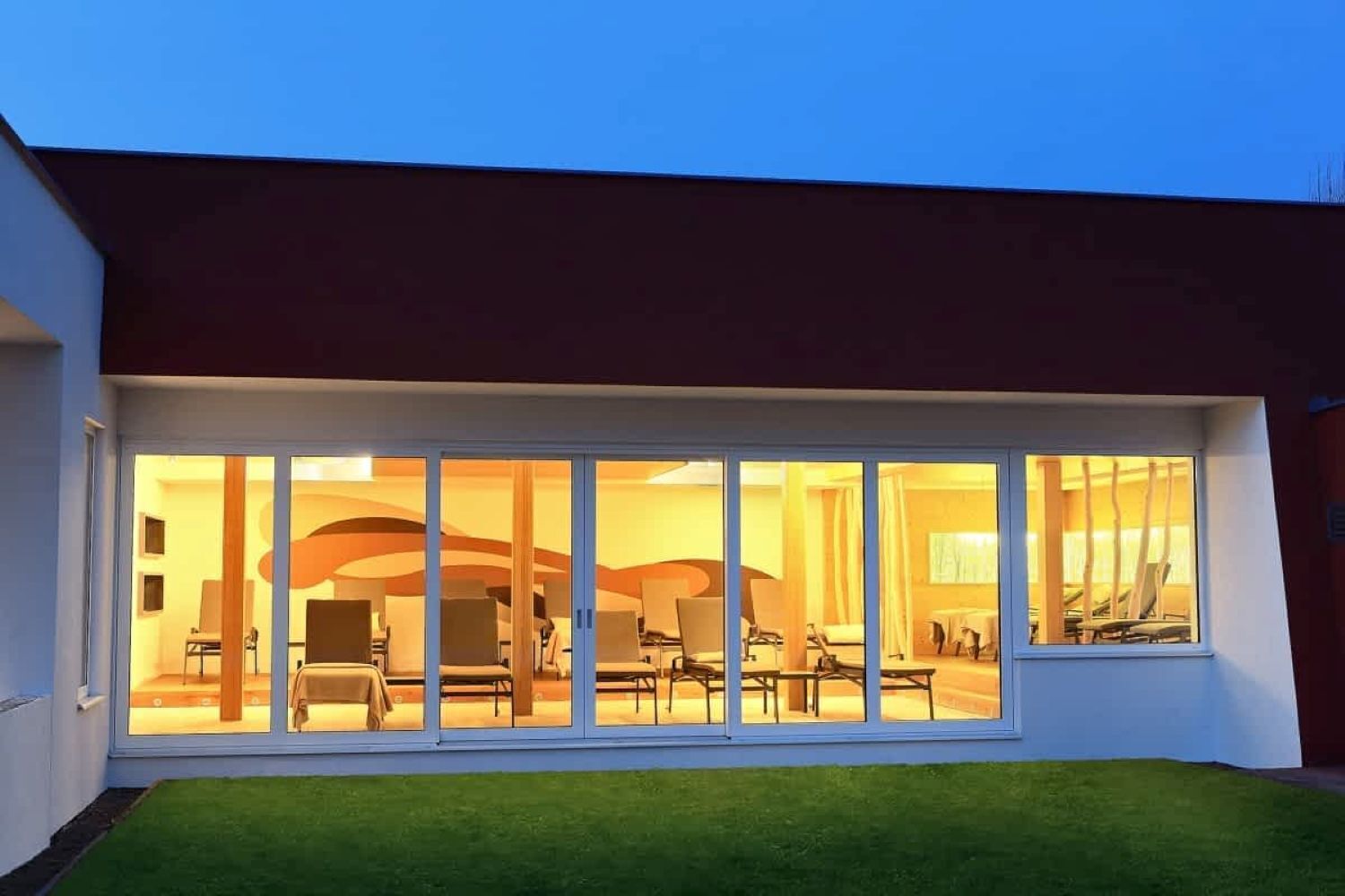 A modern house with large glass windows, a well-maintained lawn, and a visible interior with furniture.