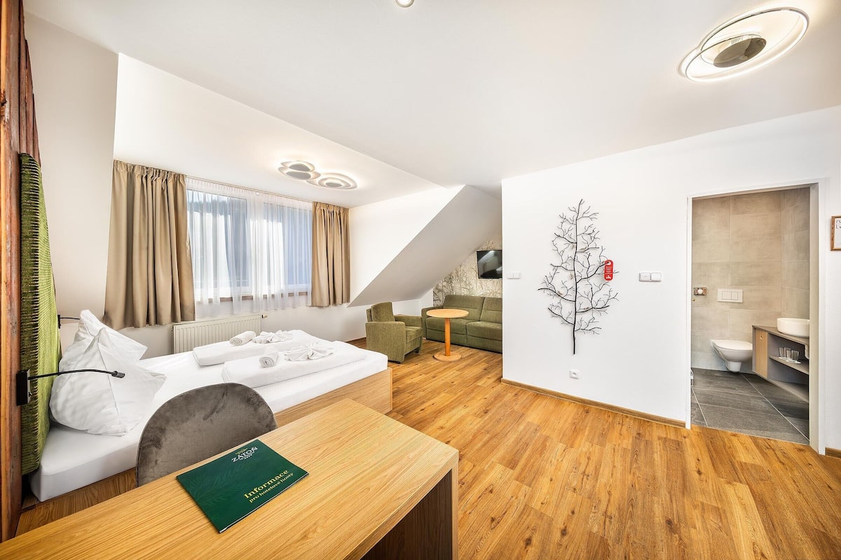 A modern hotel room with a bed, a small table, a sofa, a wall-mounted TV, and a bathroom visible through an open door.