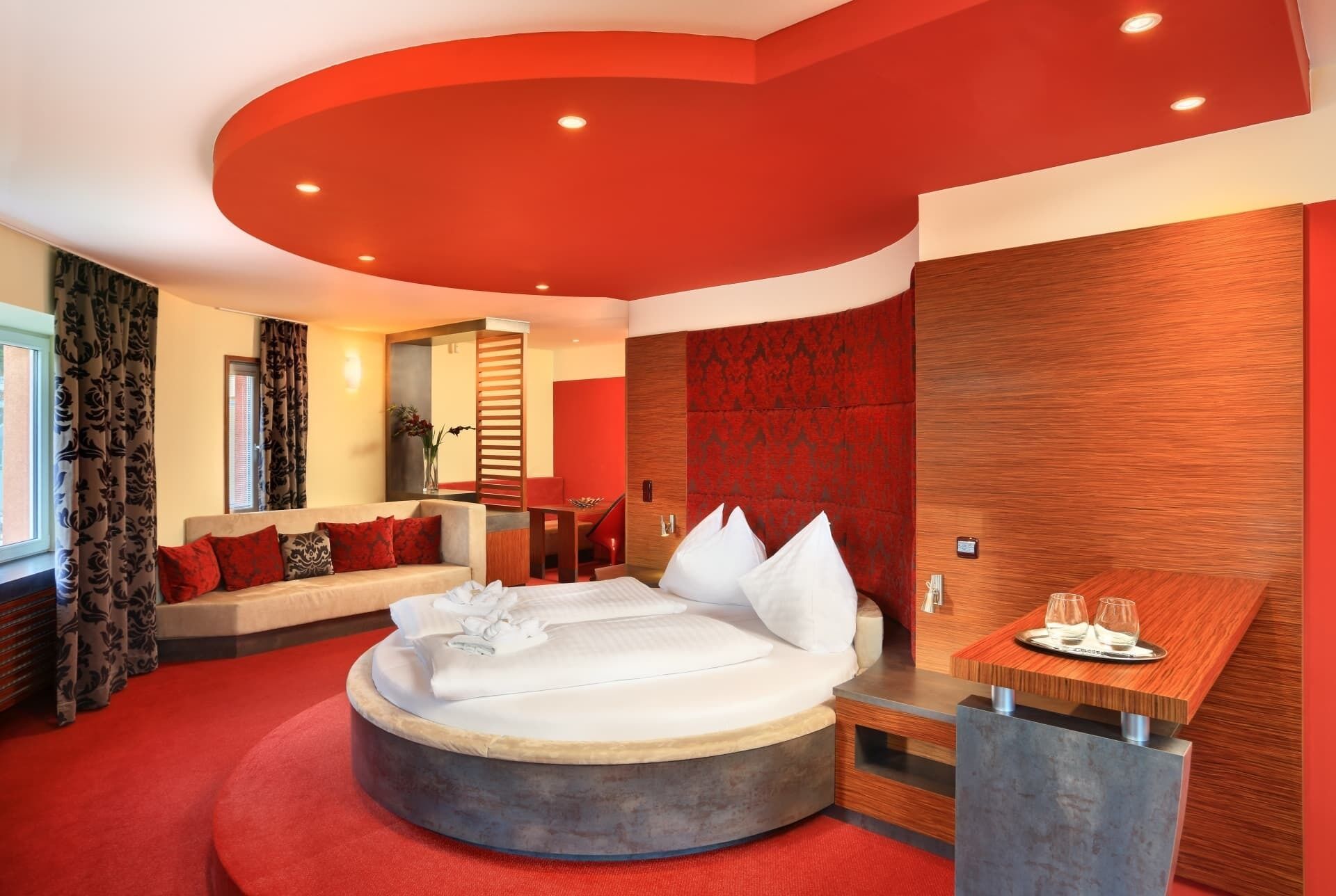 Una habitación de hotel moderna con una cama redonda, una mesita de madera, un área de descanso con sofá y una pared de acento roja.