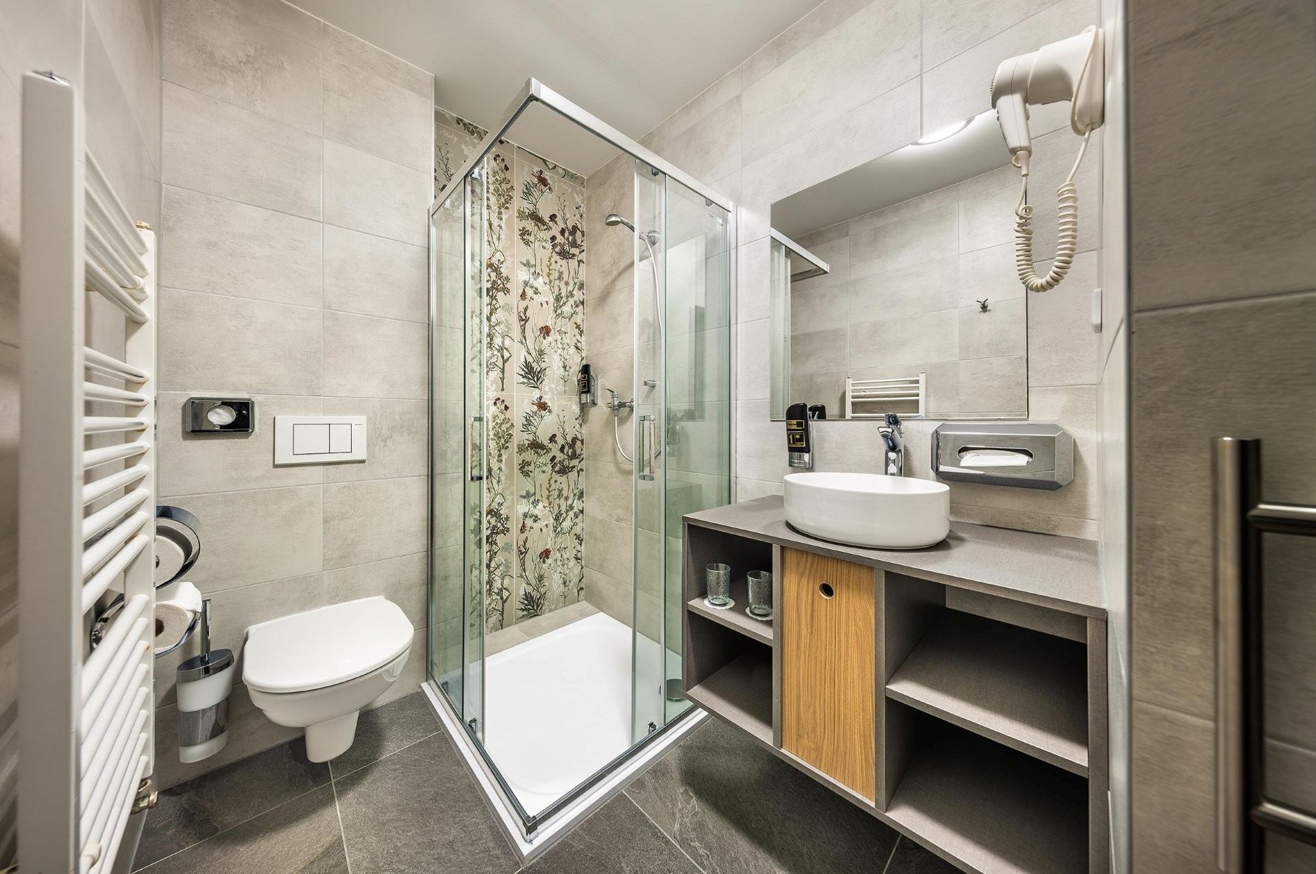Ein modernes Badezimmer mit Duschkabine aus Glas, Toilette, Waschbecken mit Handtuchspender und einem an der Wand befestigten Föhn.