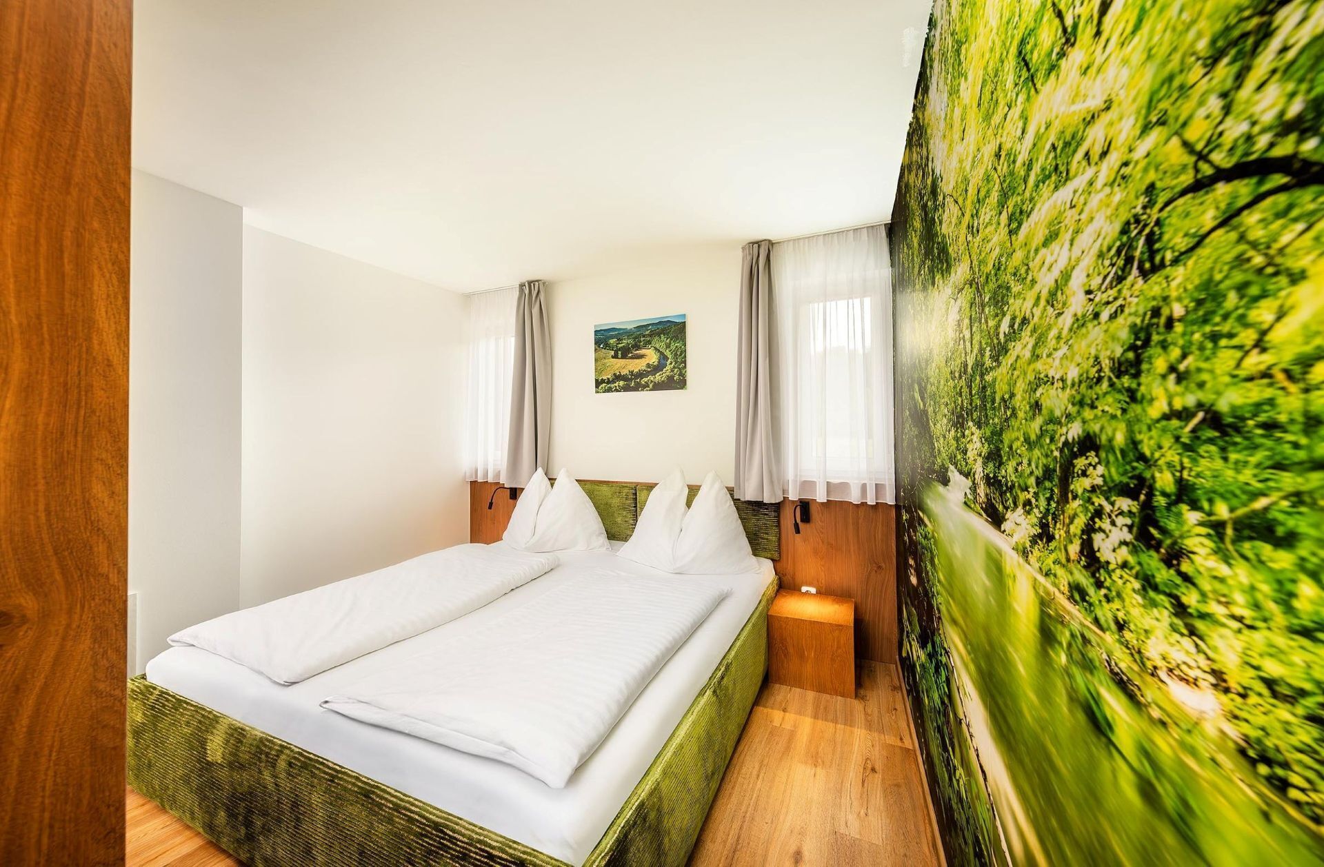A double bed with white linens, a small bedside table, a nightstand, a painting of a landscape, and a large green wall mural.