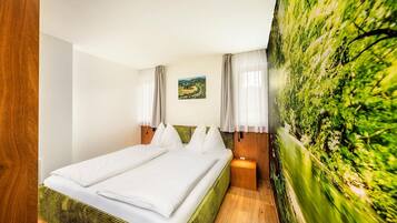 A double bed with white linens, a small bedside table, a nightstand, a painting of a landscape, and a large green wall mural.