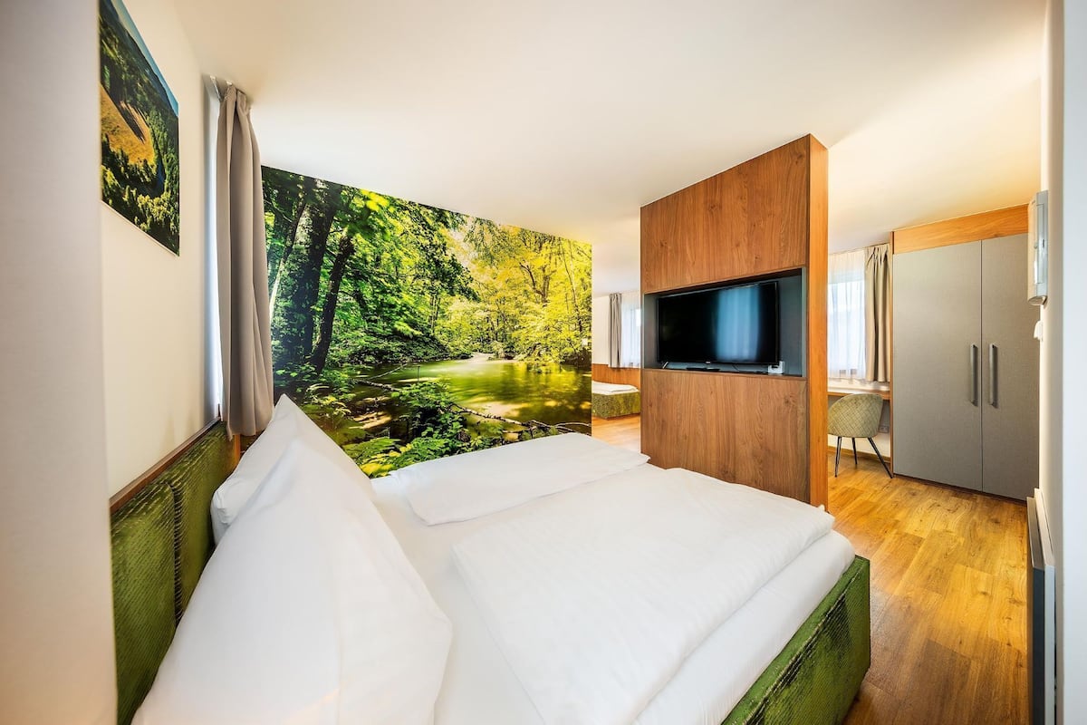 A hotel room with a bed, a TV, a desk, and a large forest mural on the wall.