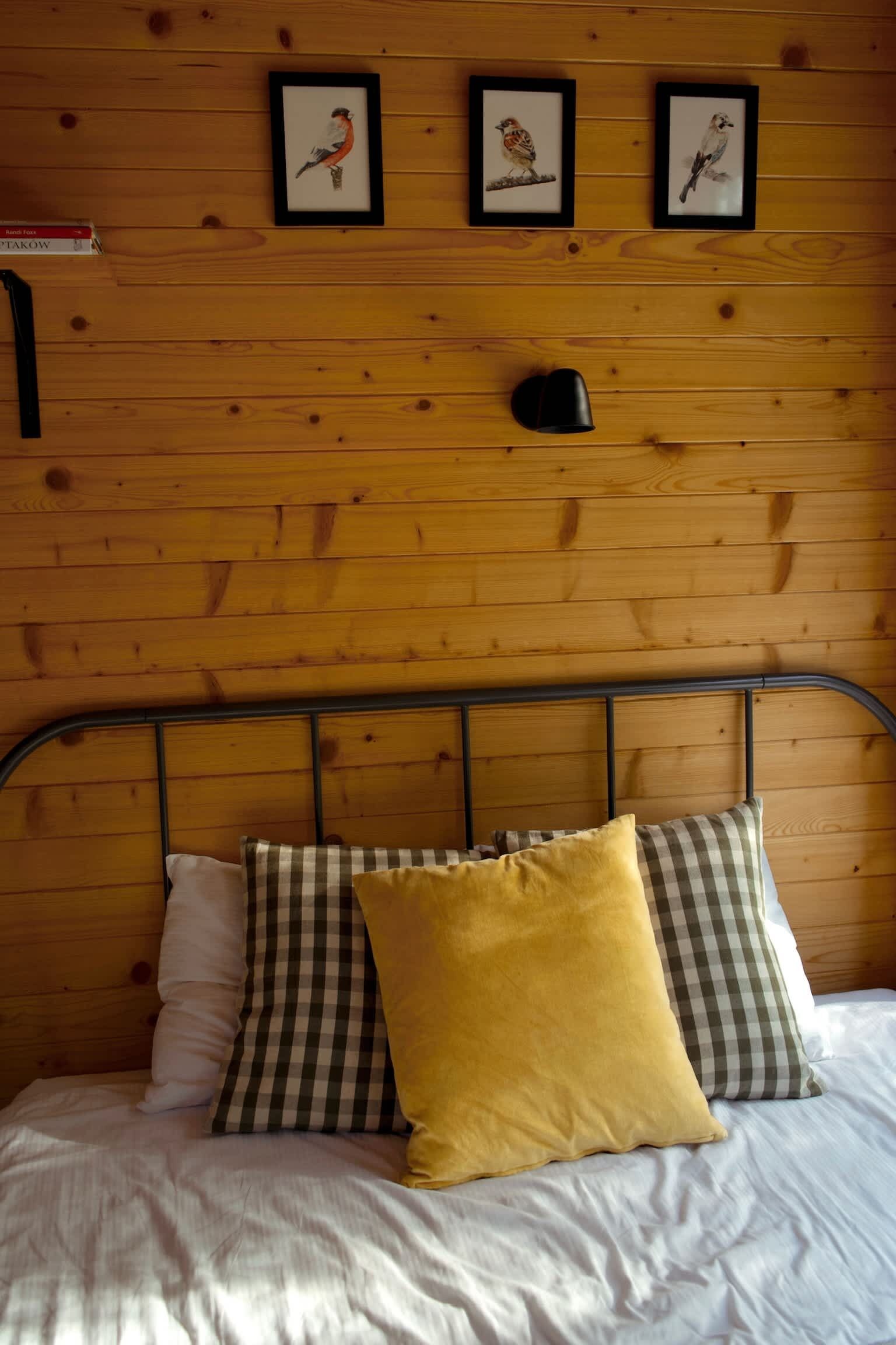 Uma cama com estrutura metálica, um travesseiro xadrez, um travesseiro amarelo e um travesseiro branco. Três quadros estão pendurados na parede acima da cama.