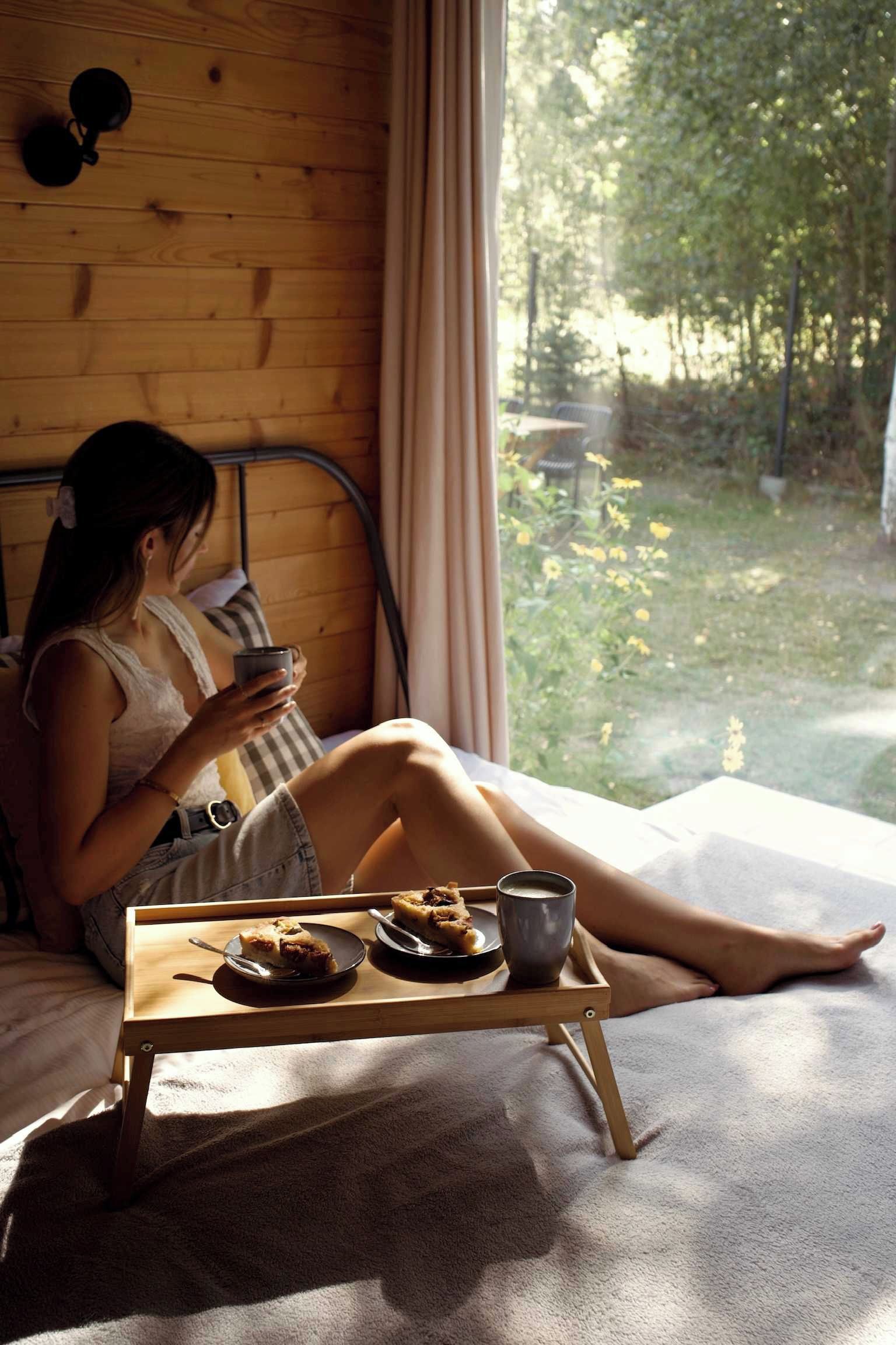 A person sitting by a window with a tray of food and a cup, in a room with wooden walls and a view of greenery outside.