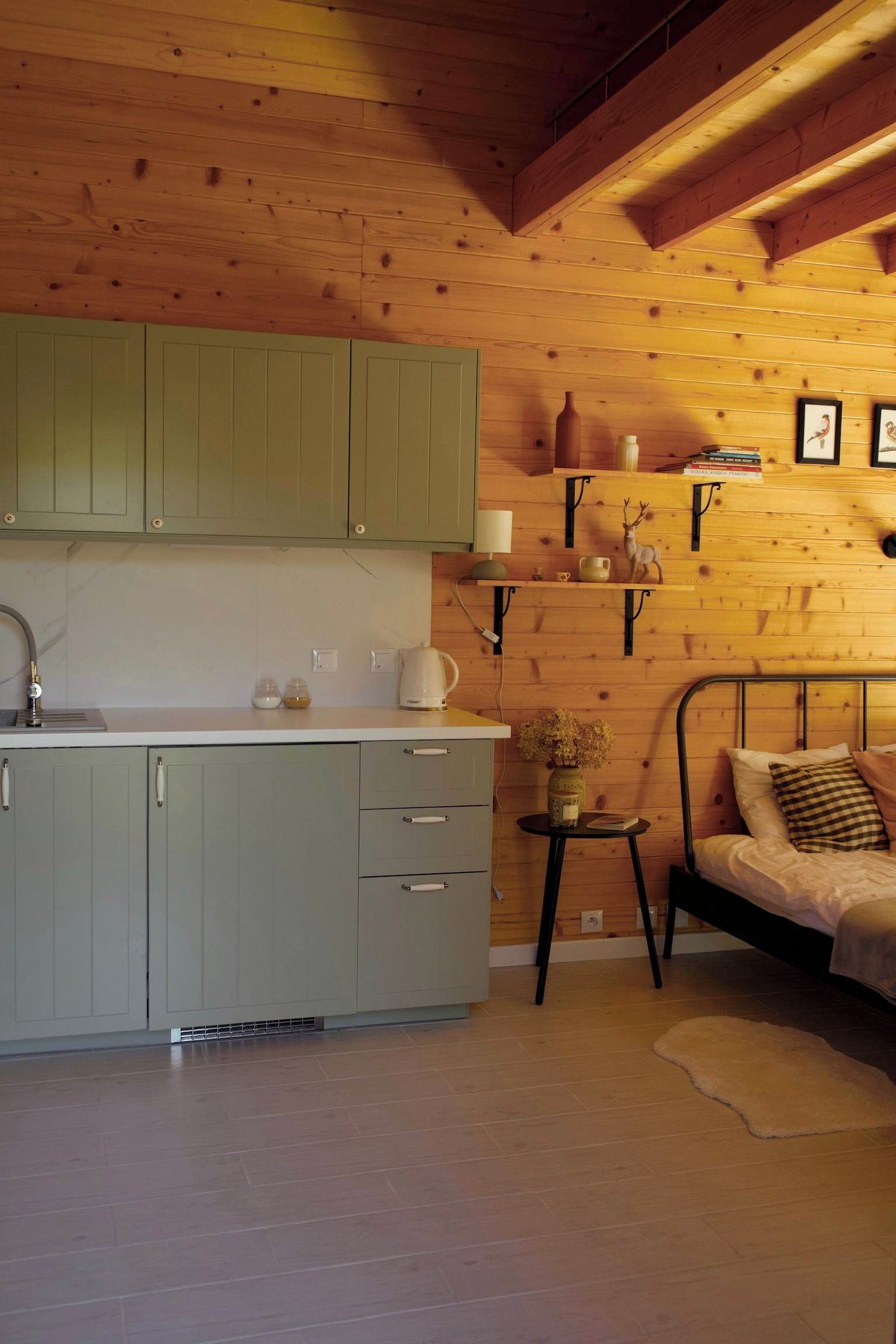 Quarto acolhedor com cama, uma mesinha com um vaso e uma cozinha com armários verdes e pia.