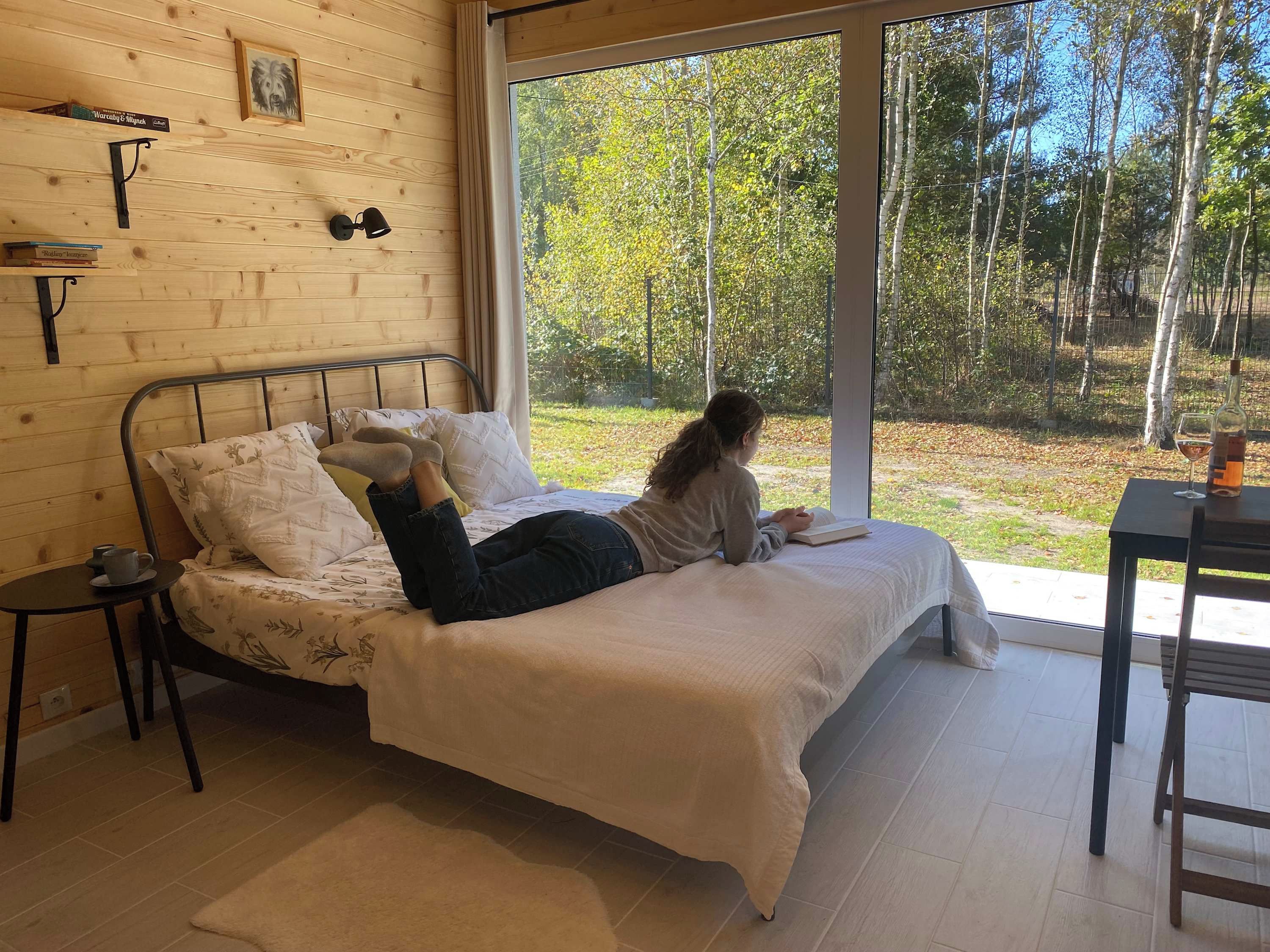 Une chambre avec un lit, une table de chevet sur laquelle se trouve une tasse, une fenêtre donnant sur des arbres et un mur orné d’un tableau et de livres.
