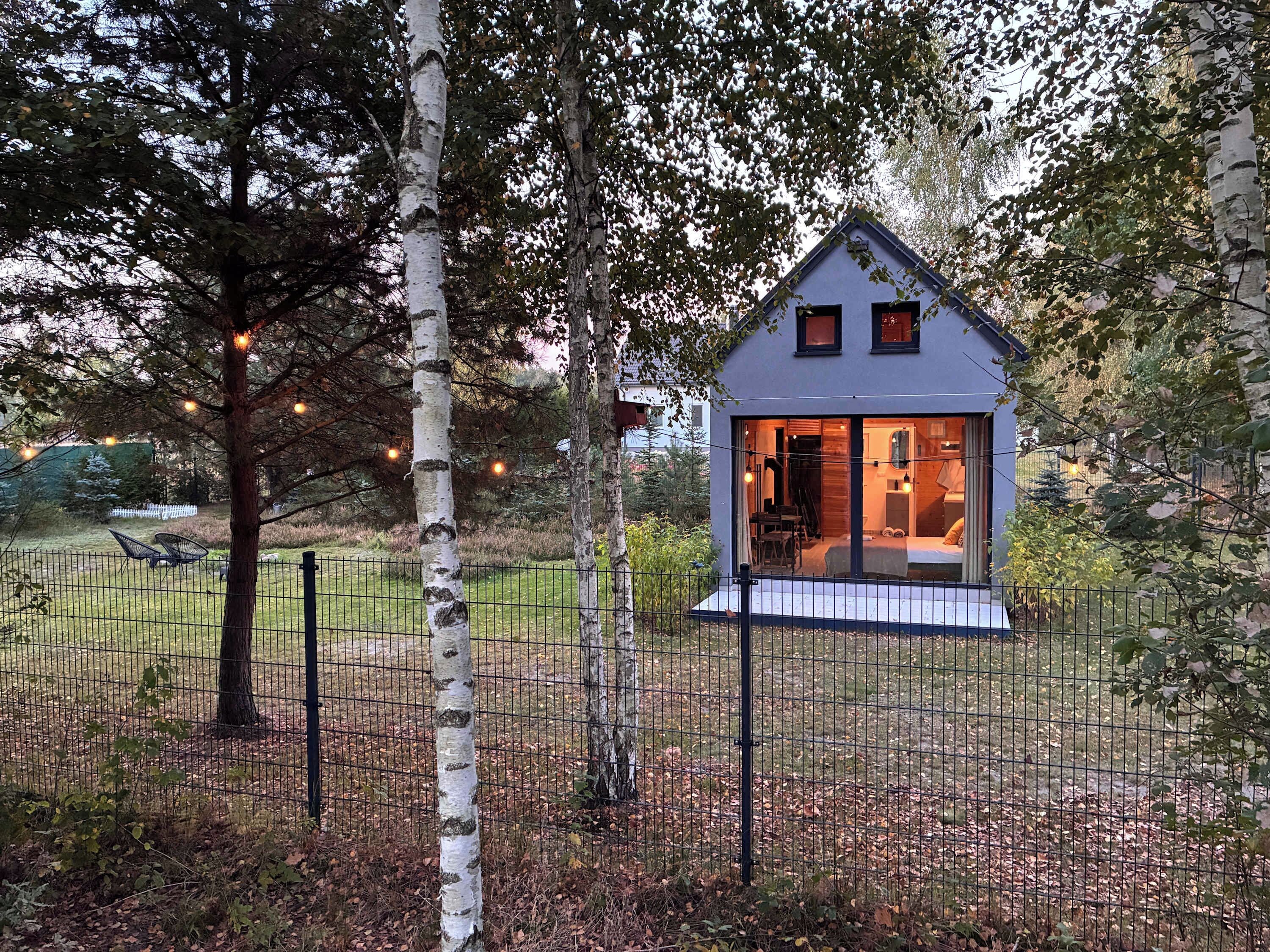 Una piccola casa moderna con interni visibili, circondata da un recinto e alberi.