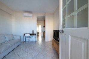 Un salón con sofá, una mesita con una silla, televisión y una cocina al fondo.