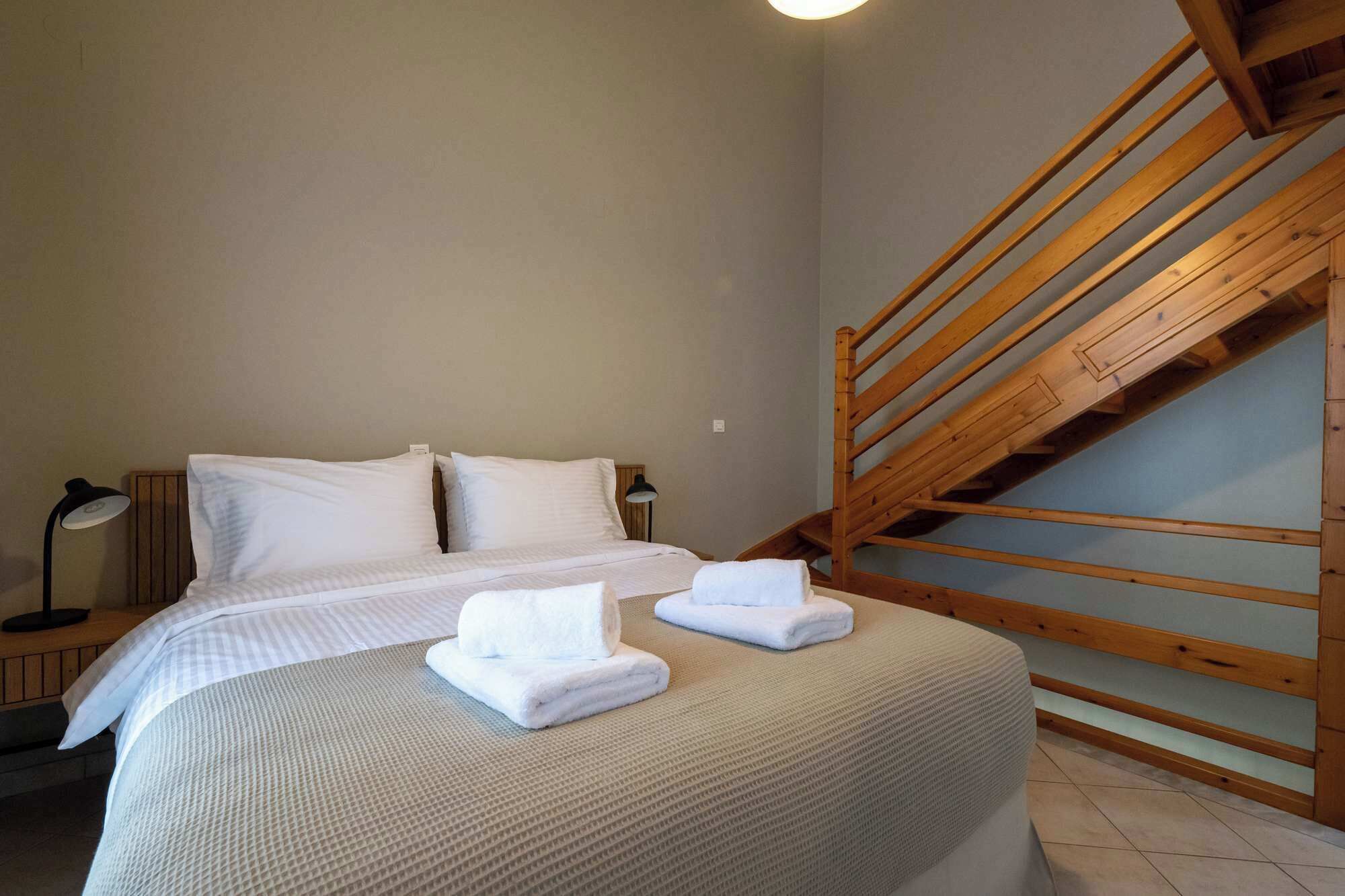 A bedroom with a bed, two pillows, a bedside lamp, a wooden staircase, and a small table.