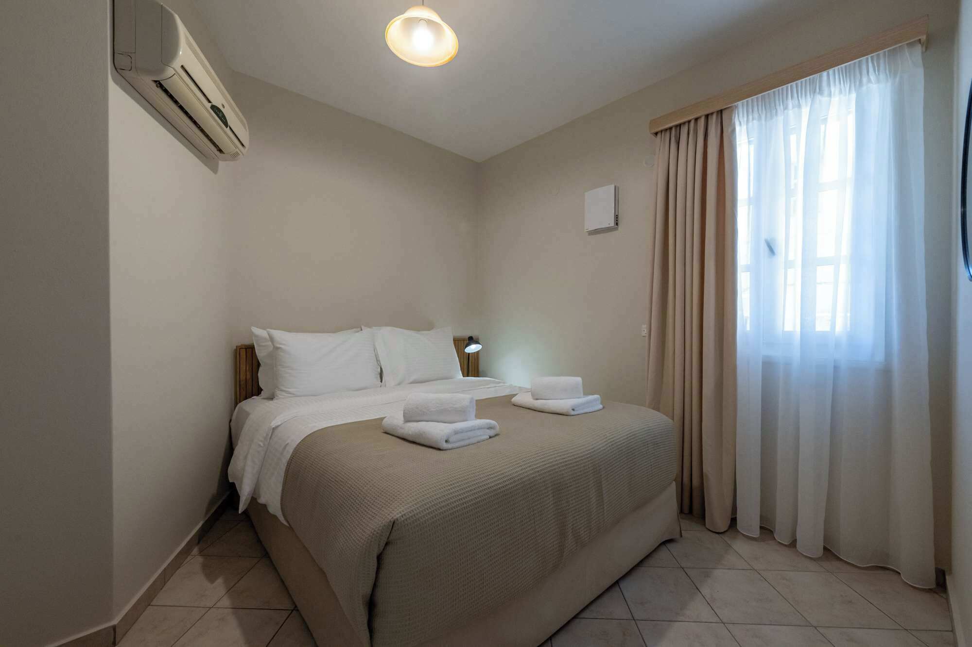 A hotel room with a bed, a bedside lamp, an air conditioning unit, and a window with curtains.