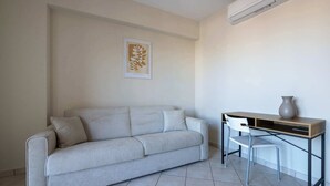 A room with a light-colored sofa, a desk with a chair, and an air conditioning unit on the wall.
