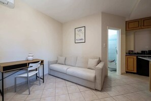 Una sala de estar con un sofá, un escritorio y una silla, un cuadro en la pared y una cocina visible a través de una puerta abierta.