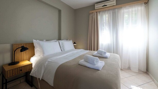 A hotel room with a bed, bedside table, lamp, air conditioning unit, and sheer curtains.
