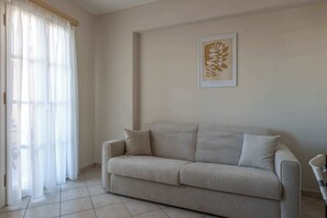 Un salón con un sofá color beige, una obra de arte enmarcada en la pared y una ventana con cortinas traslúcidas.