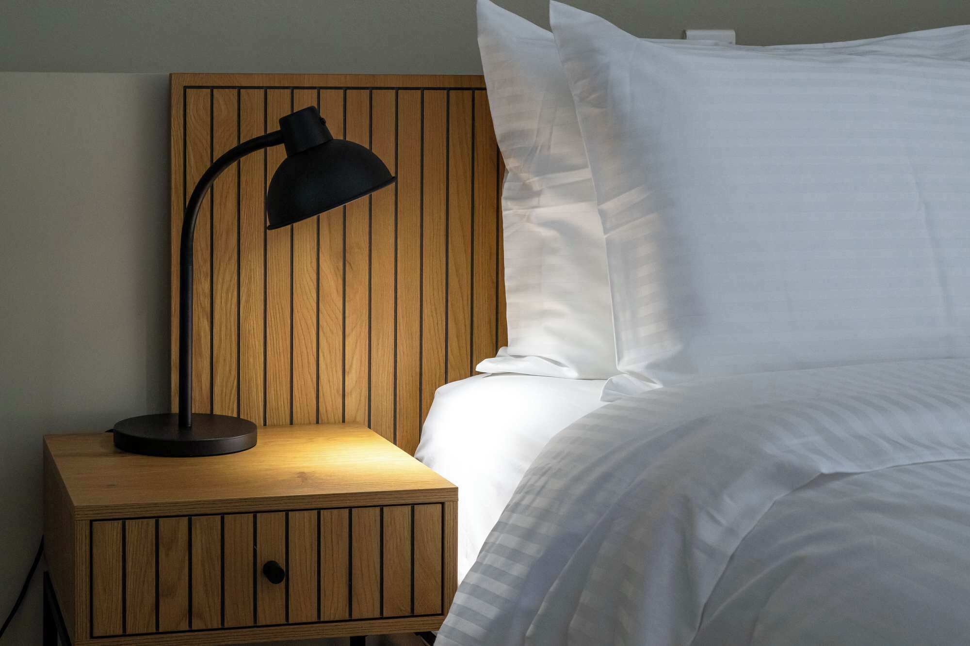 A wooden bedside table with a lamp, a bed with white bedding, and a wooden headboard.