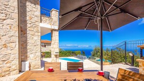 A wooden table with two mugs, a red cup, and a blue cup, an umbrella, and a stone building with a pool and a view of the sea. - Sunset Vista Villas Deluxe III (Zakynthos)