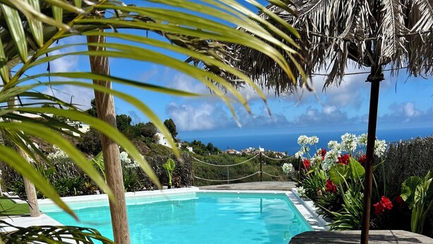 A pool area with a view of the ocean, surrounded by tropical plants and a wooden table with chairs. - Boho Villa in Firgas | Pool, Nature, BBQ, and Ocean Views (CN)