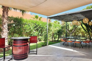 An outdoor patio with a round wooden table and chairs, a pergola with hanging lights, and a view of a garden with various plants and trees. - Villa Loulou, heated pool and breakfast included (Marrakech)