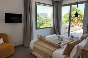 A bedroom with a bed, a TV, a window with curtains, a hanging light fixture, and a view of greenery outside. - Villa Loulou, heated pool and breakfast included (Marrakech)