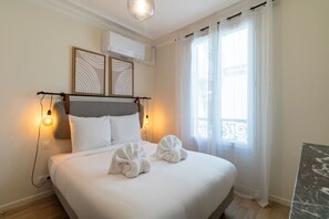 Ein ordentlich bezogenes Bett mit zwei Handtüchern, einem Kopfteil mit Lampenaufsatz und einem Fenster mit Vorhängen.
