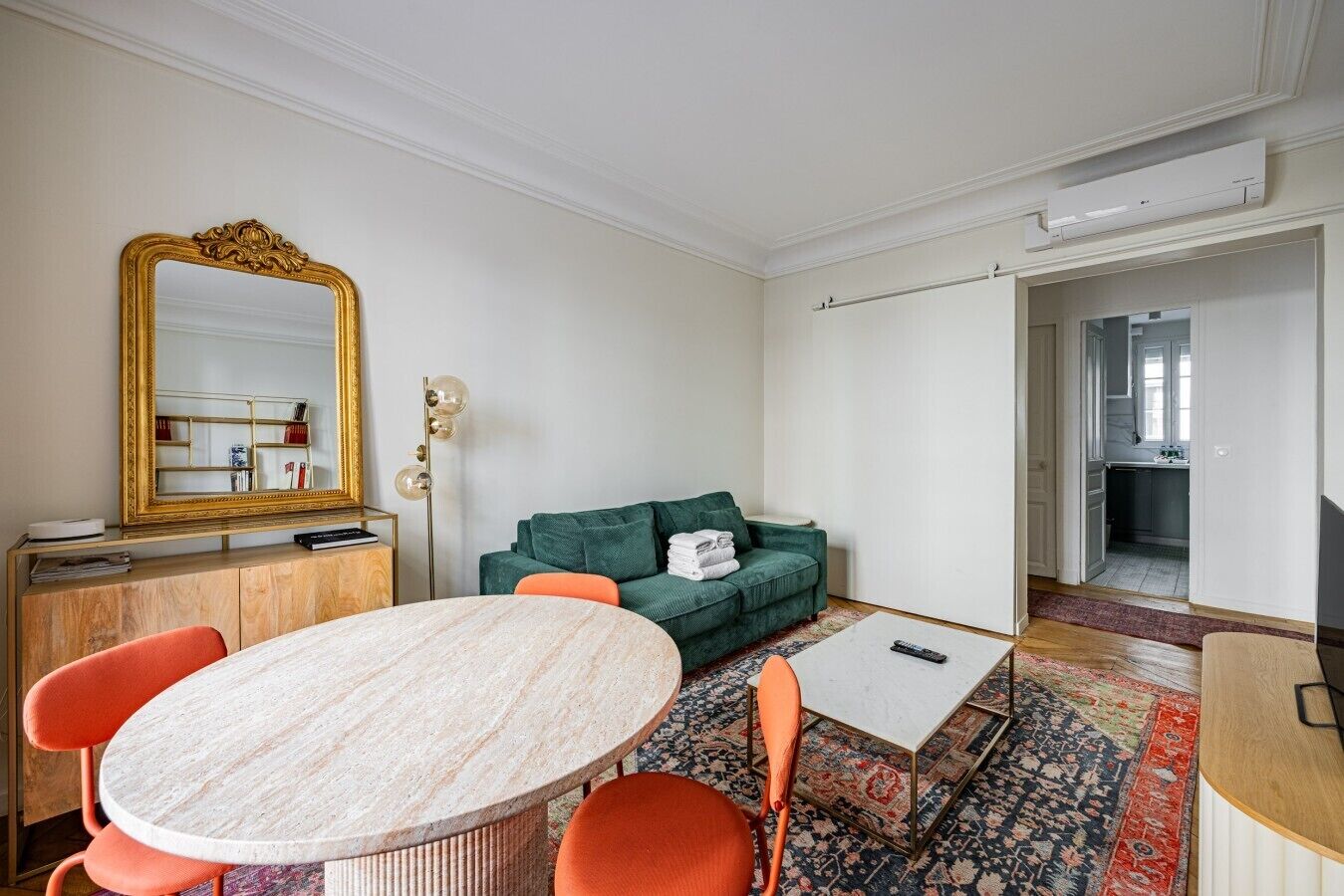 A room with a round marble table, a green sofa, a large mirror, and a patterned rug.