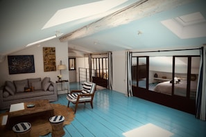Ein Loft-Zimmer mit hohen Decken, einer Couch, einem gestreiften Sessel, einem Couchtisch und Blick durch eine große Schiebetür auf ein Schlafzimmer.