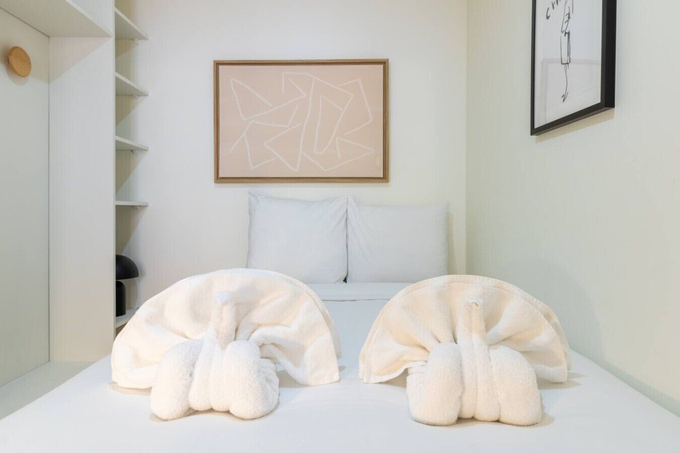 Una camera da letto con un letto, due cuscini bianchi, una parete bianca, un quadro astratto incorniciato e uno scaffale bianco con un oggetto rotondo.