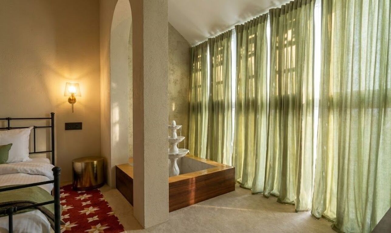 A bedroom with a bed, a bedside lamp, a wooden side table, a small round stool, and a window with sheer curtains.