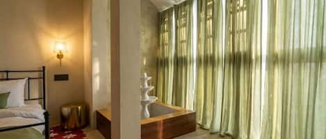 A bedroom with a bed, a bedside lamp, a wooden side table, a small round stool, and a window with sheer curtains.