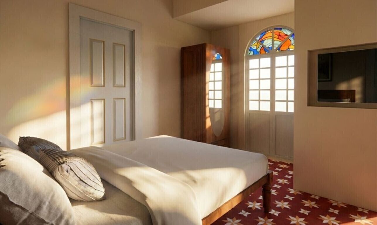 A bedroom with a bed, a wooden wardrobe, a window with a stained glass design, and a mirror on the wall.