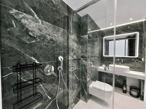 A modern bathroom with a glass shower enclosure, a white sink, and a marble-tiled wall. - Galleria Romvis – V by ΜΡS (Athina)