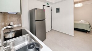 Una cocina compacta con refrigerador de acero inoxidable, un lavamanos con hervidor y una cama pequeña con colcha verde.