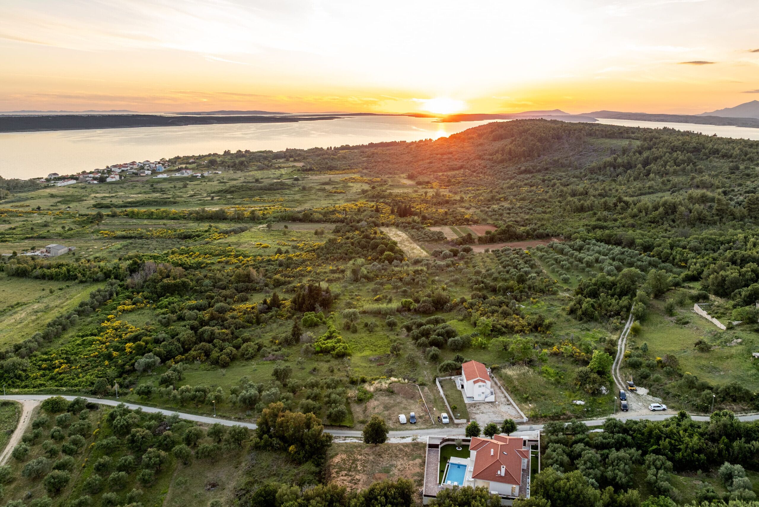 Vista aérea de una zona residencial con piscina, una carretera y un atardecer sobre una masa de agua.