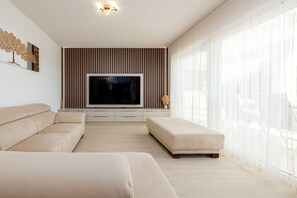 Ein modernes Wohnzimmer mit Flachbildfernseher, einem beigen Sofa, einem passenden Hocker und einem großen Fenster mit transparenten Vorhängen.