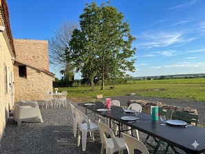 Un espace de restauration en plein air avec une table dressée pour quatre personnes, entouré de chaises blanches, d’un bâtiment en pierre et d’un grand arbre.