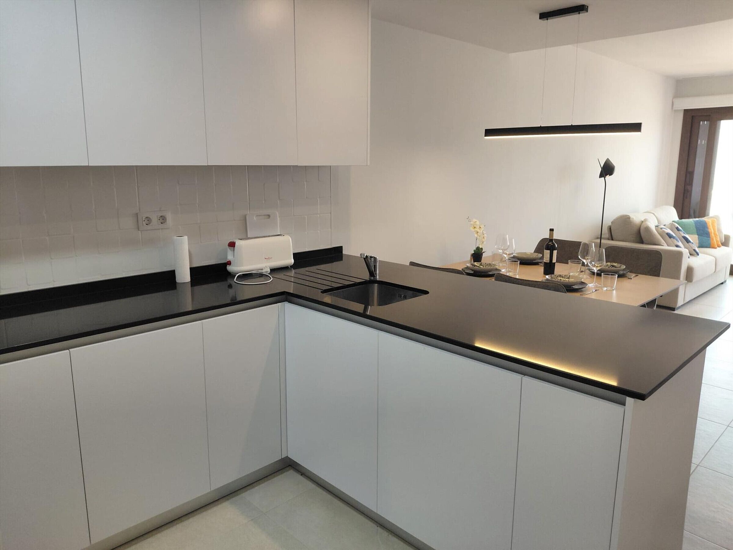 A modern kitchen with white cabinets, a black countertop, a built-in oven, and a dining area with a sofa and table.