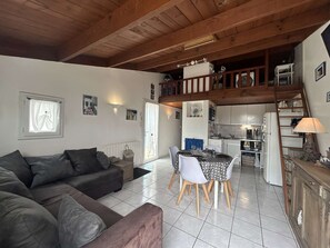 A cozy living room with a sofa, a dining table with chairs, a kitchen area with a fridge, and a staircase leading to an upper level. - Quiet house, heated pool - 5 guests (Bretignolles-sur-Mer)