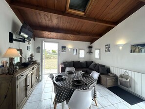 A dining area with a table set for four, a sofa, and a television mounted on the wall. - Quiet house, heated pool - 5 guests (Bretignolles-sur-Mer)