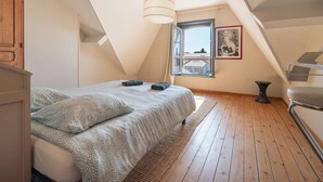 Ein Schlafzimmer mit schrägen Wänden, einem Bett mit gemusterten Betttextilien, einer Hängeleuchte, einem Fenster mit Aussicht und einem gerahmten Bild an der Wand.