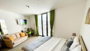 Ein Hotelzimmer mit Bett, einer Sitzecke, einem Schreibtisch, einem Fernseher und einem Balkon mit grünen Vorhängen.