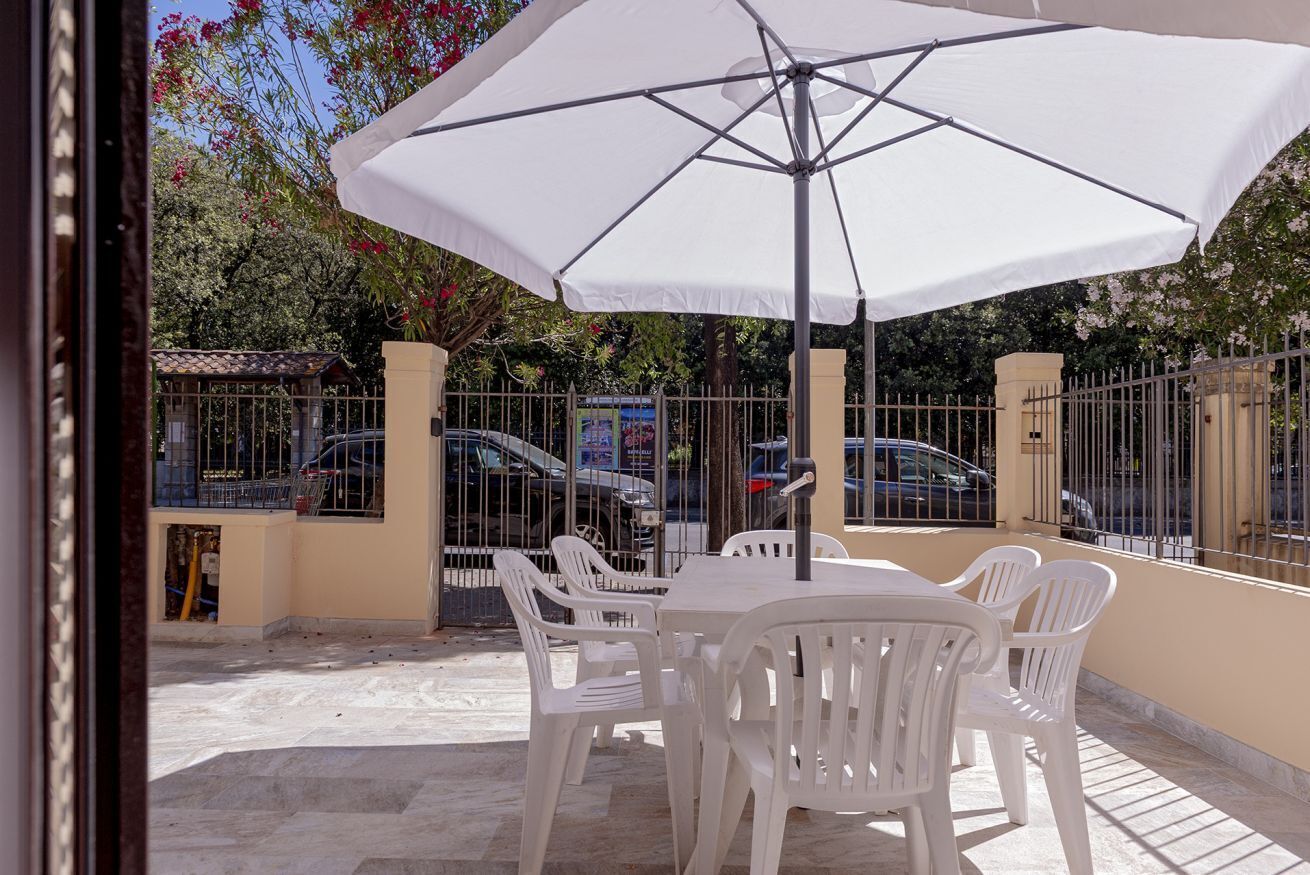 Een terras buiten met een tafel en stoelen onder een witte parasol, omringd door een metalen hek en geparkeerde auto's.